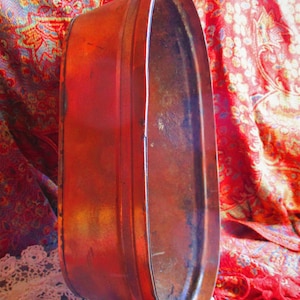 May include: A large, oval-shaped copper tub with a worn, reddish-brown patina. The tub has a slightly dented appearance and is sitting on a patterned fabric.