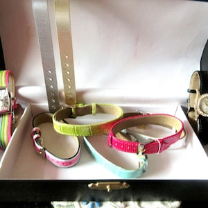Könnte beinhalten: Eine Sammlung von Uhrenarmbändern in verschiedenen Farben und Materialien, darunter Silber, Gold, Pink, Grün und Blau. Die Armbänder werden in einer schwarzen Box mit weißem Innenraum präsentiert.