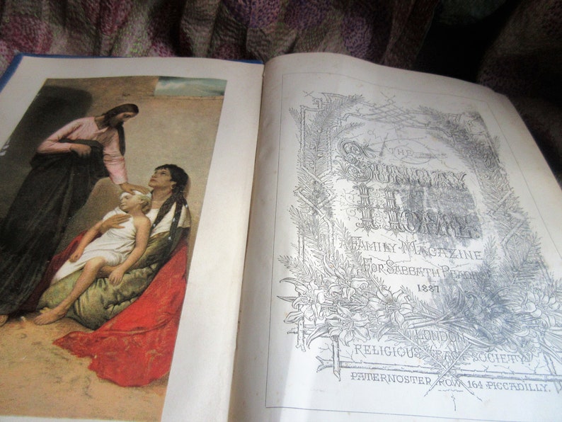 May include: An open book with a black and white illustration on the right page and a color illustration on the left page. The right page has the title 'The Sunday at Home' and the text 'Family Magazine for Sabbath Peace 1887' and 'Religious Tract Society Paternoster Row 164 Piccadilly.' The left page has a color illustration of a woman in a red dress holding a young child in a white dress. A man in a pink and black robe is standing behind the woman and touching the child's head.