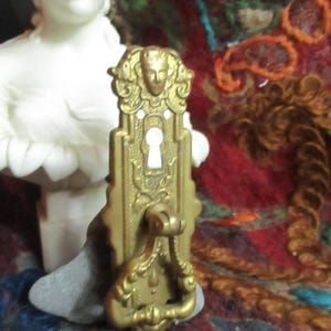 Peut inclure: Une poignée de porte en laiton vintage avec un trou de serrure et un design orné. La poignée présente un visage au centre et des détails floraux complexes.