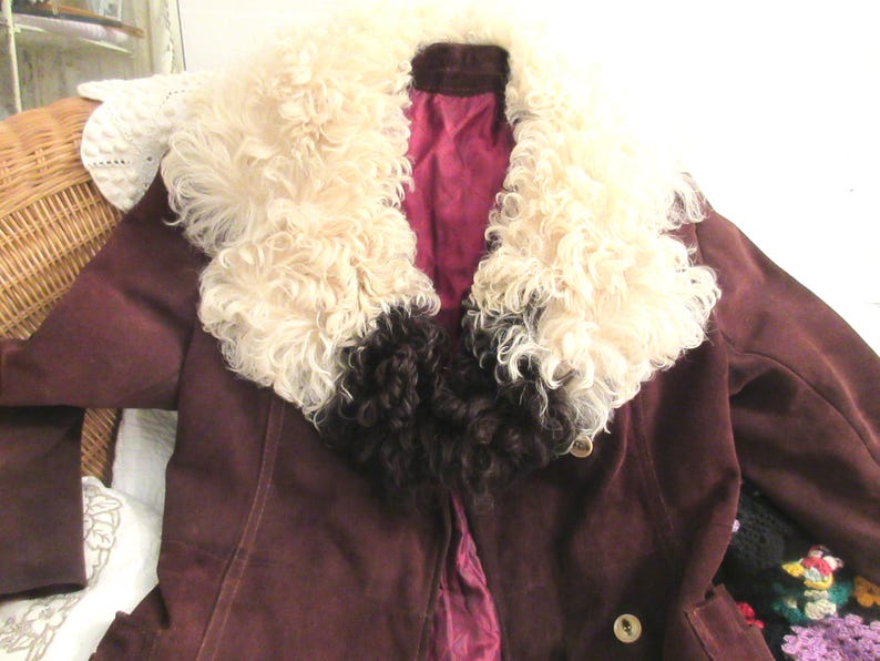 May include: A brown suede jacket with a large white and brown fur collar. The jacket has a button closure and a burgundy lining.
