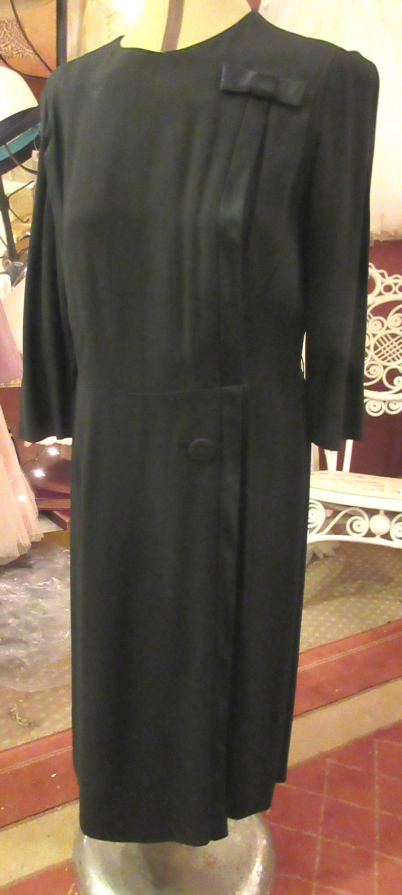 May include: A black dress with a long sleeve and a bow detail on the shoulder. The dress has a single button closure at the waist.