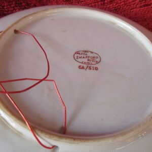 May include: White ceramic plate with a red string attached to the rim. The back of the plate has a red stamp that reads "Made in Japan, Casa Shafford Ware, CA/S10".