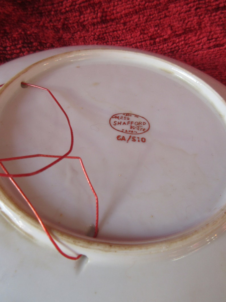 May include: White ceramic plate with a red string attached to the rim. The back of the plate has a red stamp that reads "Made in Japan, Casa Shafford Ware, CA/S10".
