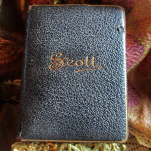 Puede incluir: Un libro encuadernado en cuero negro con letras doradas que dicen "Scott". El libro está sobre un soporte de metal dorado.