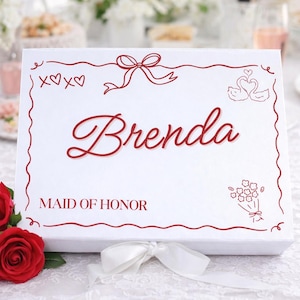 以下が含まれることがあります： 赤い装飾が施された白いギフトボックス。リボン、ハート、白鳥のイラストが描かれています。「Brenda」という名前がエレガントな筆記体で書かれ、その下に「MAID OF HONOR」と書かれています。前景には3つの赤いバラがあります。