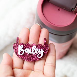 May include: A sparkly pink acrylic name tag with the name "Bailey" in white script. The tag is held in front of a pink and brown travel mug. The tag has a scalloped edge and two small holes for attachment.
