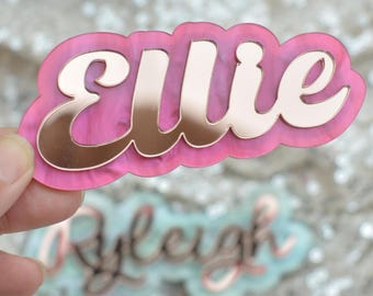 May include: A custom name sign featuring the name Ellie in a reflective rose gold script font layered over a pink marbled acrylic backing. The sign has a smooth, rounded, cloud-like shape that highlights the contrast between the metallic lettering.
