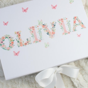 May include: White gift box with the name "OLIVIA" spelled out in floral letters. Pink butterflies and a white ribbon bow decorate the box. The box is on a white, textured surface.
