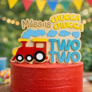May include: A red frosted cake with a wooden train cake topper. The topper has the name "Miles is" and the words "Chugga Chugga" and "Two Two" in blue and yellow. The cake sits on a wooden cake stand.