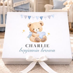 May include: A white gift box with a watercolor illustration of a teddy bear holding a blue heart. The box is tied with a white ribbon. The name "Charlie Benjamin Brown" is written on the box in blue script. A stuffed teddy bear and a crib are in the background.
