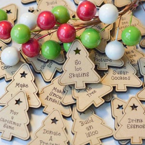 K&ouml;nnte beinhalten: Ein Stapel h&ouml;lzerner Weihnachtsbaumschmuck mit verschiedenen Aktivit&auml;ten darauf geschrieben, wie "Schlittschuhlaufen gehen", "Schneengel machen", "Ein Weihnachts-Spiel spielen" und "Ein Geschenk spenden".