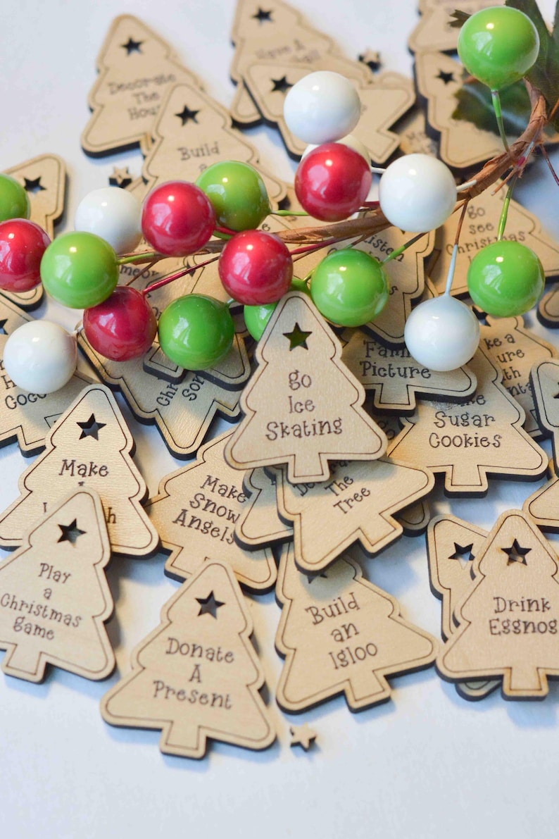 K&ouml;nnte beinhalten: Ein Stapel h&ouml;lzerner Weihnachtsbaumschmuck mit verschiedenen Aktivit&auml;ten darauf geschrieben, wie "Schlittschuhlaufen gehen", "Schneengel machen", "Ein Weihnachts-Spiel spielen" und "Ein Geschenk spenden".