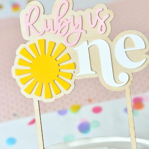 May include: A wooden cake topper featuring the text Ruby is one in light pink and white lettering. A bright yellow sun icon is positioned between the words. The topper is mounted on two thin wooden sticks and is inserted into a white frosted cake.