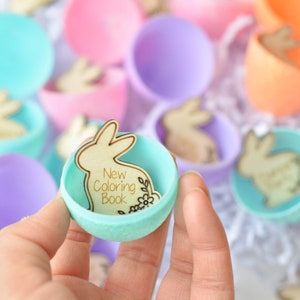 K&ouml;nnte beinhalten: Ein hellblaues Plastik-Osterei mit einem ausgeschnittenen Holzhasen im Inneren. Der Hase tr&auml;gt die Aufschrift "New Coloring Book".