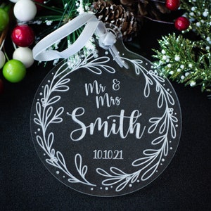 May include: Clear acrylic Christmas ornament with the text "Mr & Mrs Smith 10.10.21" surrounded by a floral wreath design. The ornament has a white ribbon for hanging. The background includes greenery and berries.