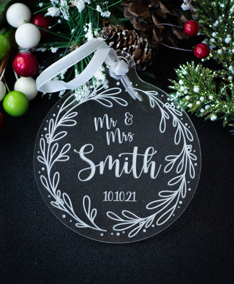 May include: Clear acrylic Christmas ornament with the text "Mr & Mrs Smith 10.10.21" surrounded by a floral wreath design. The ornament has a white ribbon for hanging. The background includes greenery and berries.