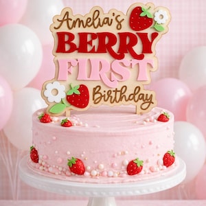 May include: A pink frosted cake decorated with strawberries and pearls, topped with a wooden sign that reads "Amelia's BERRY FIRST Birthday". The cake sits on a white cake stand, with pink and white balloons in the background.