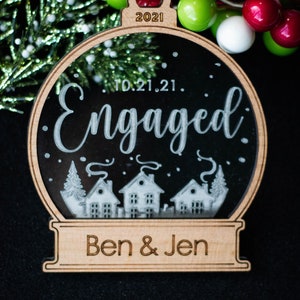 May include: Wooden Christmas ornament with a clear center, featuring the word "Engaged" and the date "10.21.21." The ornament has a small town scene at the bottom and the names "Ben & Jen". The top of the ornament has the year "2021".