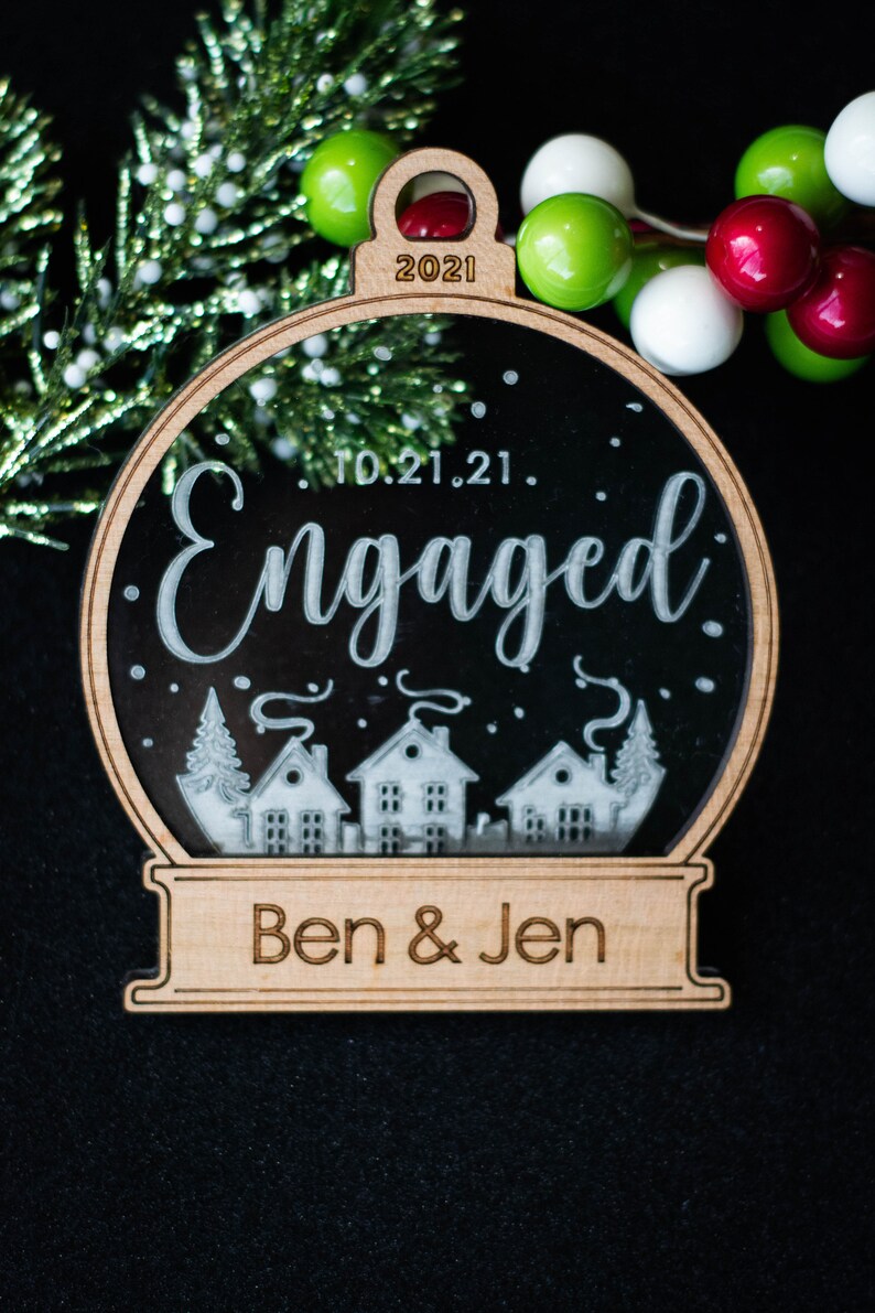 May include: Wooden Christmas ornament with a clear center, featuring the word "Engaged" and the date "10.21.21." The ornament has a small town scene at the bottom and the names "Ben & Jen". The top of the ornament has the year "2021".