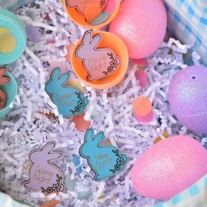 May include: An Easter basket filled with colorful eggs and decorative bunny-shaped tags. The tags have phrases like "Movie Night" and "New Outfit". The basket is lined with white paper and has a blue and white checkered pattern.