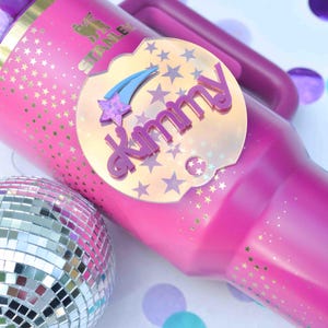 Könnte beinhalten: Ein rosafarbener Edelstahlbecher mit einem goldenen Sternenmuster und einem rosa- und schillernden Aufkleber mit der Aufschrift "dreamy" mit einem Sternschnuppen- und Sternenmotiv. Der Becher steht neben einer silbernen Discokugel.