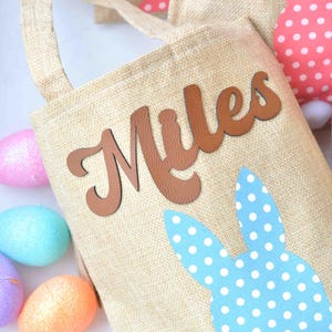 May include: A beige burlap Easter basket with the name "Miles" in brown letters. The basket has a blue bunny ear design with white polka dots. Colorful Easter eggs are scattered around the basket.
