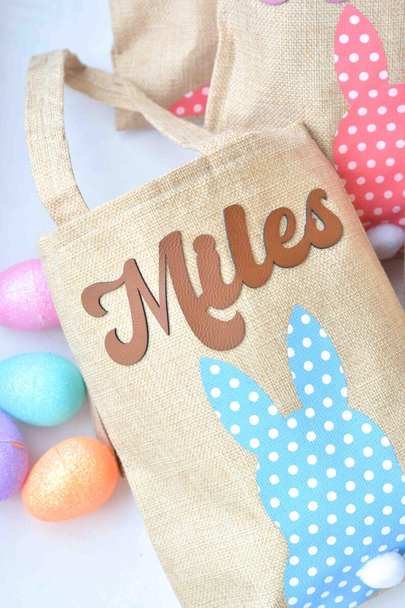 May include: A beige burlap Easter basket with the name "Miles" in brown letters. The basket has a blue bunny ear design with white polka dots. Colorful Easter eggs are scattered around the basket.