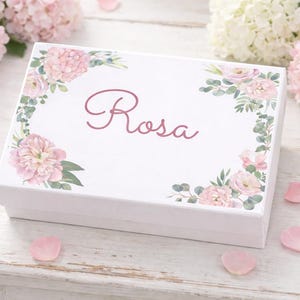 May include: A rectangular white gift box featuring a floral design with pink peonies, green eucalyptus leaves, and the name Rosa written in a dark pink cursive script on the lid. The box is decorated with a botanical pattern at the corners.