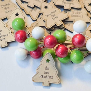 K&ouml;nnte beinhalten: Eine Sammlung von h&ouml;lzernen Weihnachtsbaumschmuck mit verschiedenen Aktivit&auml;ten, die darauf geschrieben sind, wie "Drink Hot Coco", "Decorate The Tree" und "Go Ice Skating". Der Schmuck ist auf einer wei&szlig;en Oberfl&auml;che mit roten, gr&uuml;nen und wei&szlig;en Dekorbeeren verstreut.