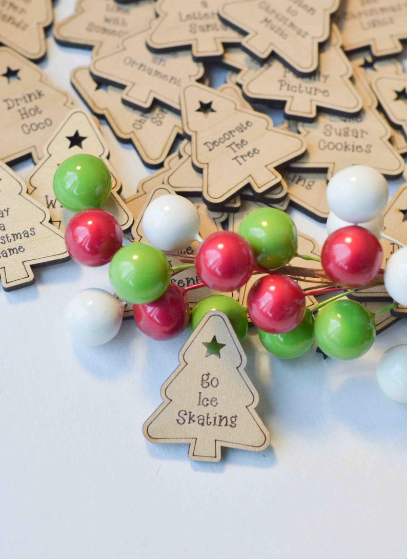 K&ouml;nnte beinhalten: Eine Sammlung von h&ouml;lzernen Weihnachtsbaumschmuck mit verschiedenen Aktivit&auml;ten, die darauf geschrieben sind, wie "Drink Hot Coco", "Decorate The Tree" und "Go Ice Skating". Der Schmuck ist auf einer wei&szlig;en Oberfl&auml;che mit roten, gr&uuml;nen und wei&szlig;en Dekorbeeren verstreut.