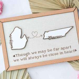 May include: A wooden sign with a light brown frame and a beige linen background. The sign features two state outlines, Michigan and Tennessee, with a heart and arrow connecting them. The text reads "Though we may be far apart we will always be close in heart."