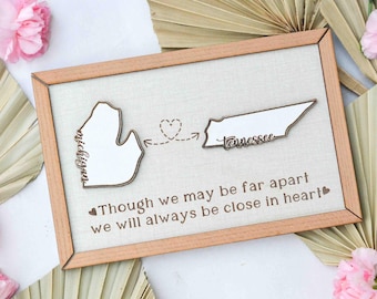 May include: A wooden sign with a light brown frame and a beige linen background. The sign features two state outlines, Michigan and Tennessee, with a heart and arrow connecting them. The text reads "Though we may be far apart we will always be close in heart."