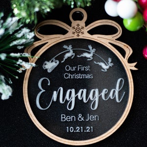 May include: A wooden Christmas ornament with a black center and a light brown border. The ornament reads "Our First Christmas Engaged Ben & Jen 10.21.21" with a reindeer and sleigh design. The ornament is shaped like a Christmas ball.