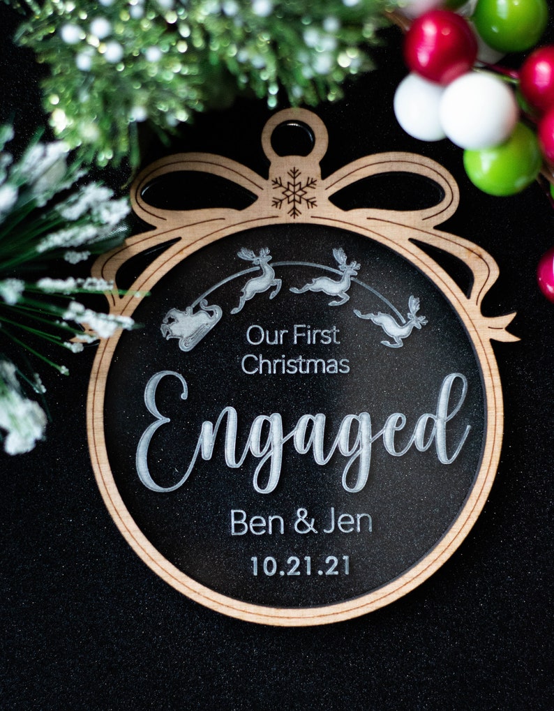 May include: A wooden Christmas ornament with a black center and a light brown border. The ornament reads "Our First Christmas Engaged Ben & Jen 10.21.21" with a reindeer and sleigh design. The ornament is shaped like a Christmas ball.