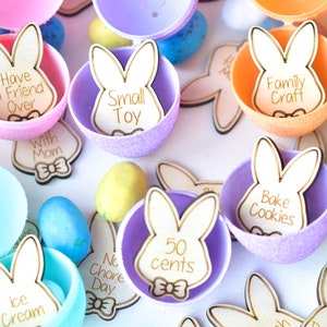 May include: Easter-themed image featuring colorful plastic eggs with glitter and wooden bunny-shaped tags. The tags have phrases like "Small Toy," "Family Craft," and "50 cents" engraved on them. Other Easter eggs are scattered around.
