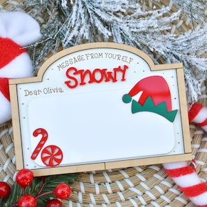 Puede incluir: Un letrero de madera con el texto "MESSAGE FROM YOUR ELF" y el nombre "Snowy" en rojo. El letrero tiene un espacio en blanco para un mensaje y está decorado con un gorro de elfo rojo y verde. Bastones de caramelo y un caramelo de menta también están en la imagen.