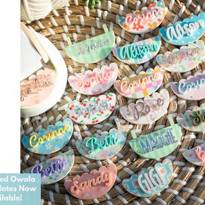 Puede incluir: Una colecci&oacute;n de placas con nombres estampados con varios nombres en escritura cursiva, incluyendo "Corinne" y "Allison". Las placas tienen bordes festoneados y est&aacute;n dispuestas sobre una superficie tejida. El texto "Patterned Owala Name Plates Now Available!" es visible.
