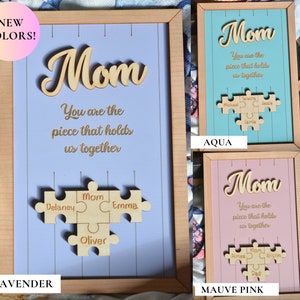 Puede incluir: Letrero enmarcado de madera con la palabra "Mom" en cursiva y la frase "You are the piece that holds us together". Debajo, un rompecabezas de madera con nombres. Disponible en lavanda, aguamarina y rosa malva.