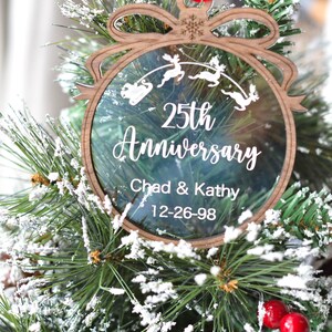 May include: A wooden Christmas ornament with a clear center, featuring the text "25th Anniversary" and the names "Chad & Kathy" with the date "12-26-98." The ornament has a reindeer and sleigh design and is hanging on a Christmas tree.