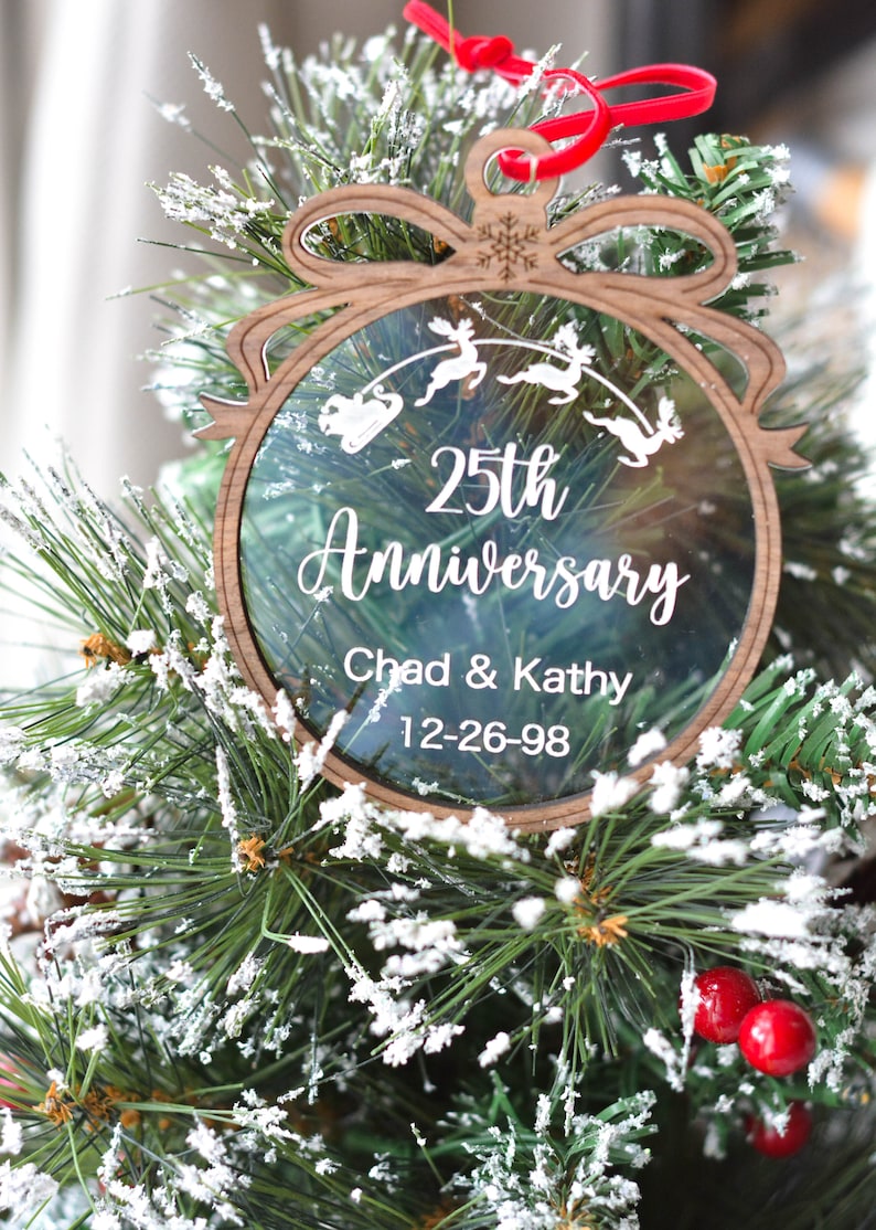 May include: A wooden Christmas ornament with a clear center, featuring the text "25th Anniversary" and the names "Chad & Kathy" with the date "12-26-98." The ornament has a reindeer and sleigh design and is hanging on a Christmas tree.