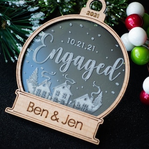 May include: A wooden Christmas ornament with a clear center, featuring the word "Engaged" and the date "10.21.21." Below the text are etched houses and trees. The ornament is personalized with "Ben & Jen" and the year "2021".