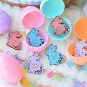 May include: An assortment of pastel-colored Easter eggs and open plastic eggshells. Inside the eggshells are bunny-shaped cards with phrases like "New Outfit," "Sweet Treat," and "Movie Night." The eggs and cards are on a white, fluffy surface.