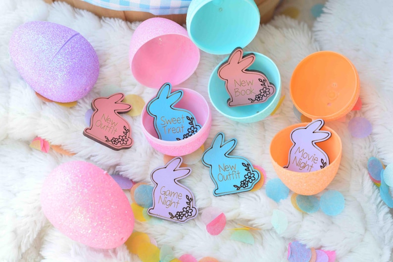 May include: An assortment of pastel-colored Easter eggs and open plastic eggshells. Inside the eggshells are bunny-shaped cards with phrases like "New Outfit," "Sweet Treat," and "Movie Night." The eggs and cards are on a white, fluffy surface.