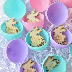 May include: Colorful Easter egg halves in shades of pink, purple, and teal, filled with wooden bunny cutouts. Each bunny has a different activity engraved on it, such as "Sweet Treat," "Movie," and "Family Craft." The eggs are arranged on white paper.