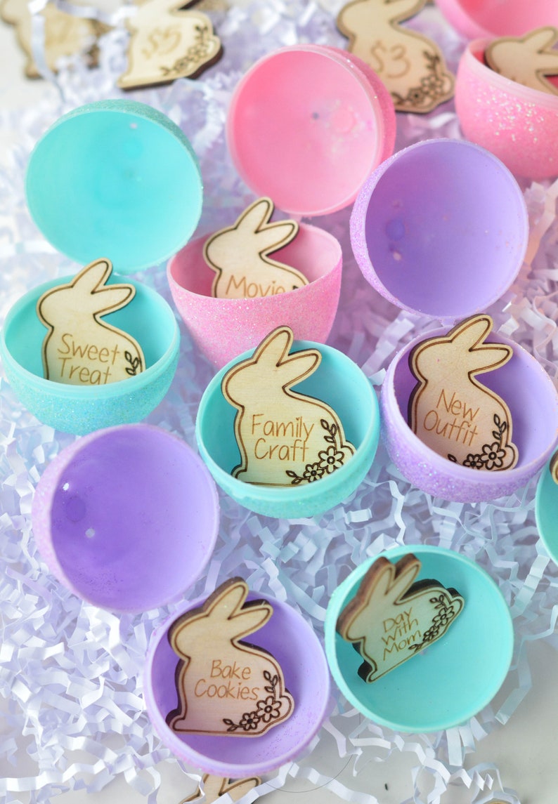 May include: Colorful Easter egg halves in shades of pink, purple, and teal, filled with wooden bunny cutouts. Each bunny has a different activity engraved on it, such as "Sweet Treat," "Movie," and "Family Craft." The eggs are arranged on white paper.