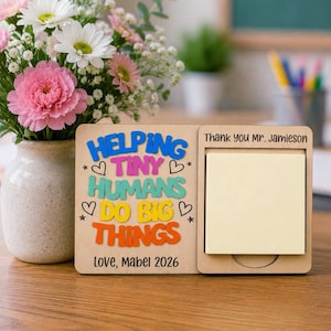 Puede incluir: Un organizador de escritorio de madera clara con un bloc de notas adhesivas amarillo cuadrado y el texto colorido "HELPING TINY HUMANS DO BIG THINGS" con "Thank you Mr. Jamieson" y "Love, Mabel 2026" grabados en la superficie frontal.