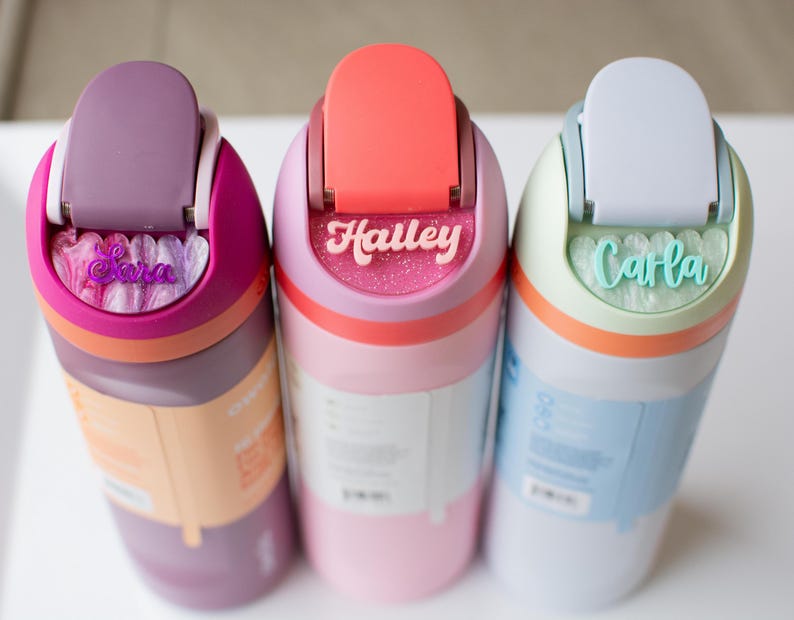 Puede incluir: Tres botellas de agua coloridas con nombres personalizados: "Cara", "Haley" y "Carla". Cada botella presenta un esquema de color diferente, incluyendo morado, rosa y azul, con tapas y detalles a juego. Los nombres est&aacute;n en cursiva.