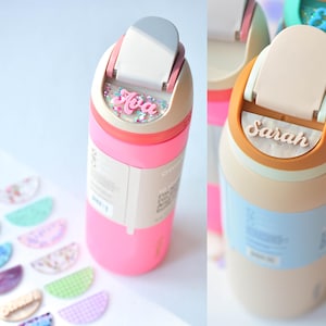 Puede incluir: Botellas de agua rosas y beige con nombres personalizados, como "Ava" y "Sarah". Las botellas tienen una tapa abatible y un dise&ntilde;o colorido. Tambi&eacute;n se ven etiquetas de nombre decorativas.