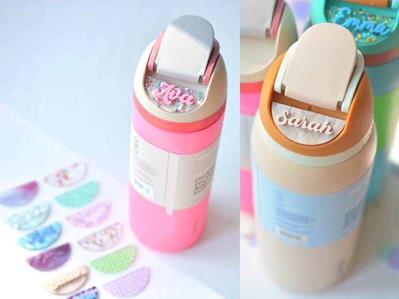 Puede incluir: Botellas de agua rosas y beige con nombres personalizados, como "Ava" y "Sarah". Las botellas tienen una tapa abatible y un dise&ntilde;o colorido. Tambi&eacute;n se ven etiquetas de nombre decorativas.